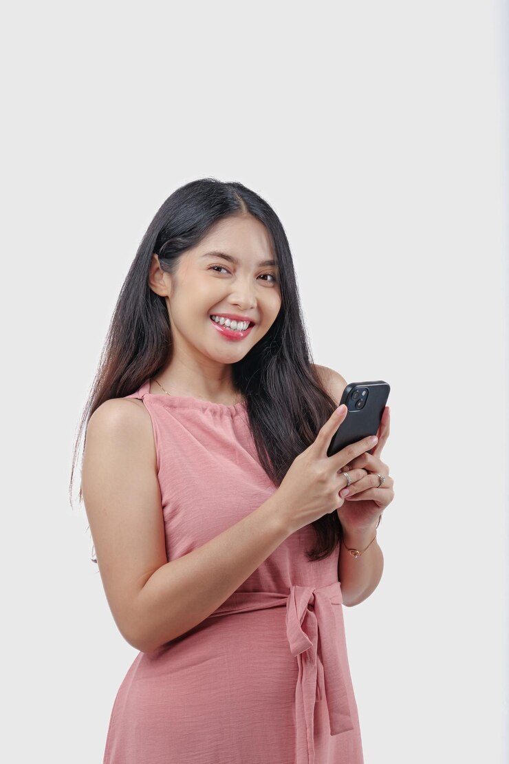 Eine fröhliche junge asiatische Frau in einem rosa Kleid hält ein Smartphone in der Hand und zeigt einen glücklichen Ausdruck