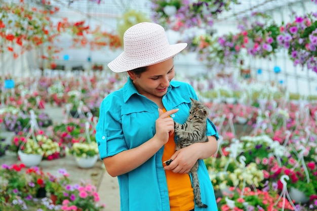 Eine fröhliche junge Dame eines Gewächshauses mit Blumen hält ein graues Kätzchen in ihren Händen