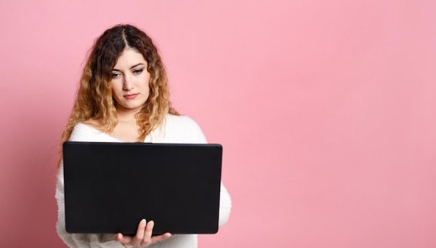 Eine fröhliche junge Frau, die auf einem rosa Hintergrund steht und einen Laptop mit einem Platz für einen Kopierbereich verwendet