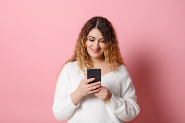 Eine fröhliche junge Frau, die isoliert auf einem rosa Hintergrund mit einem Smartphone in der Hand steht