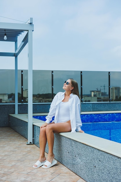 Eine fröhliche junge frau in einem weißen badeanzug und einem weißen hemd sitzt am pool eine frau mit sonnenbrille und badeanzug sommerurlaub am pool gebräuntes mädchen