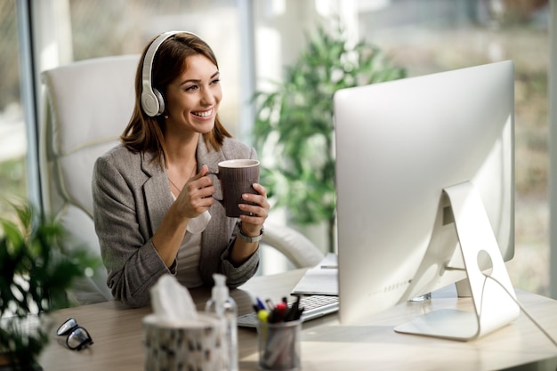 Eine fröhliche junge Frau mit Kopfhörern genießt eine Tasse Kaffee, während sie im Heimbüro am Computer arbeitet.