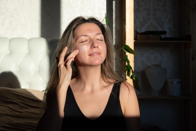 Foto eine fröhliche junge kaukasische frau lächelt mit geschlossenen augen und legt creme auf ihr gesicht.