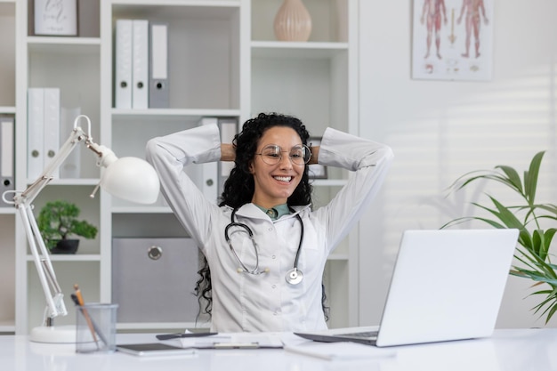 Eine fröhliche lateinamerikanische Ärztin in weißem Mantel macht eine Pause an ihrem Büro mit einem Laptop, fühlt sich zufrieden und