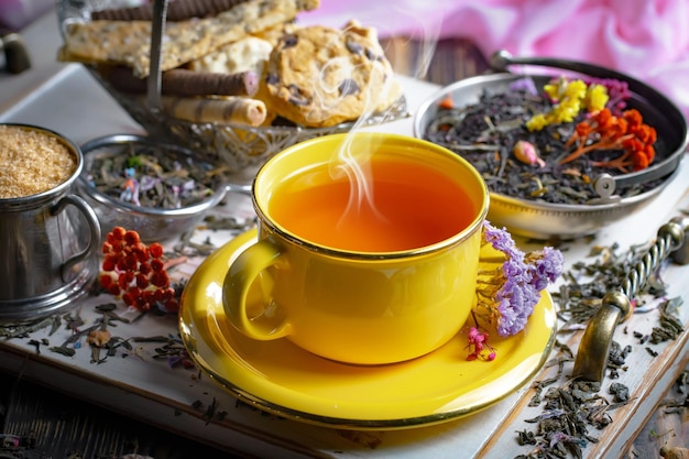 Eine gelbe Tasse Tee steht auf einem Tisch neben einem Stapel Teeblätter und Blumen.