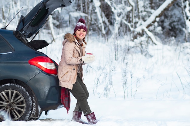 Eine gemütliche Frau lächelt, eine Frau in warmer Winterkleidung trinkt ein heißes Getränk, Tee oder Kaffee, sitzt im Kofferraum eines Autos und lächelt. Urlaub, Autofahrt, Schneekälte. Speicherplatz kopieren