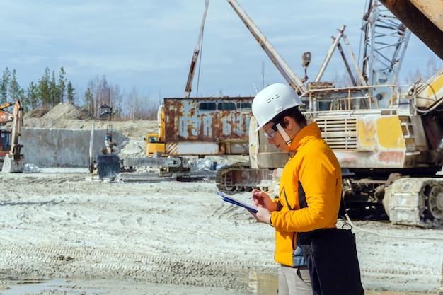 Eine Geologin oder ein Bergbauingenieur schreibt etwas in einen Kartenkoffer inmitten eines Steinbruchs mit Baumaschinen