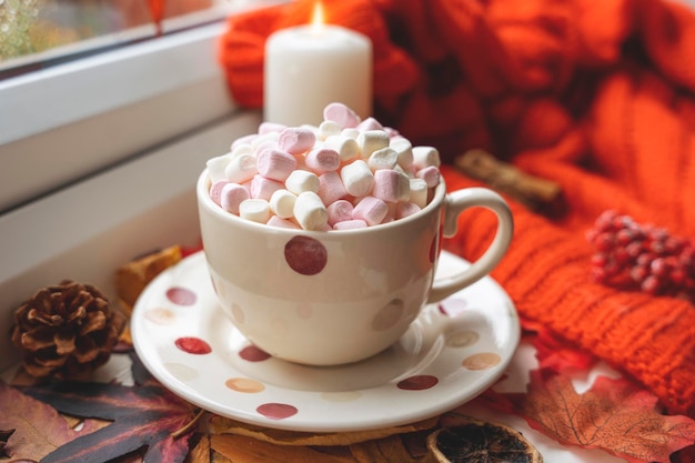 Eine gepunktete Marshmallow-Tasse mit einer Untertasse auf Herbstblättern mit einem orangefarbenen Schal, einer Kerze und einem Kegel