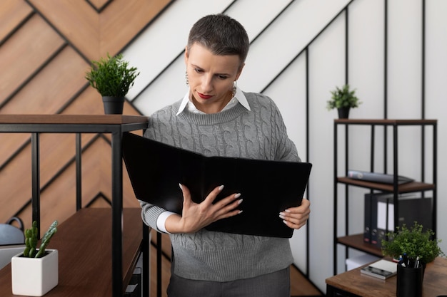Eine Geschäftsfrau mit kurzem Haarschnitt in einem grauen Pullover steht mit einer Ledermappe im Büro