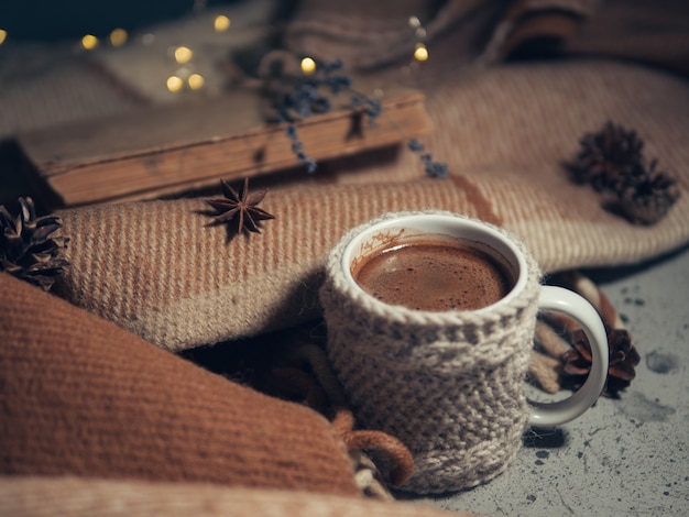 Eine gestrickte Tasse heißen Kaffees auf einer warmen Decke und ein altes Buch