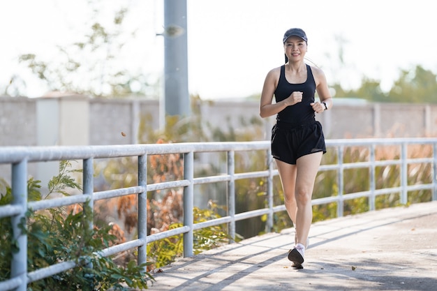 Eine gesunde glückliche asiatische Läuferin in schwarzen Sportoutfits, die im natürlichen Stadtpark unter abendlichem Sonnenuntergang joggt