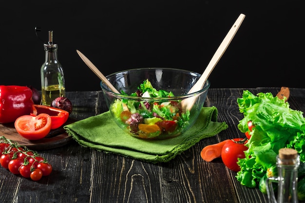 Eine Glasschüssel mit frischem Gemüsesalat, umgeben von den Zutaten: grüner Salat, Gurken, Paprika und Tomaten auf schwarzem Hintergrund