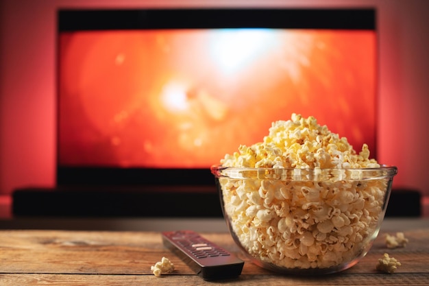 Eine Glasschüssel mit Popcorn und Fernbedienung, im Hintergrund funktioniert der Fernseher. Abends gemütlich einen Film oder eine Fernsehserie zu Hause anschauen