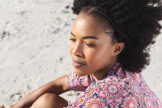 Eine glückliche afroamerikanische Frau lächelt und blickt weg, sitzend am sonnigen Strand. Sommer, Wohlbefinden, Freiheit, Entspannung und Urlaub, unverändert.