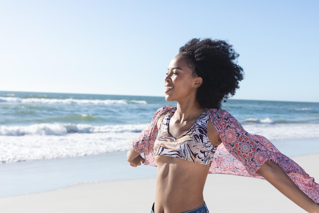 Eine glückliche afroamerikanische Frau lächelt und geht am sonnigen Strand am Meer. Sommer, Wohlbefinden, Freiheit, Entspannung und Urlaub, unverändert.