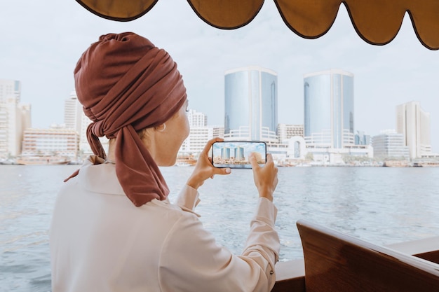 Eine glückliche Asiatin mit rotem Turban fotografiert auf ihrem Smartphone, während sie auf einem traditionellen Abra-Dhow-Boot auf dem Dubai Creek Travel in den VAE fährt