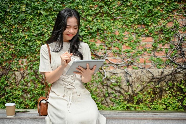 Eine glückliche asiatische Frau nutzt ihr digitales Tablet, während sie auf einer Bank in der Stadt entspannt