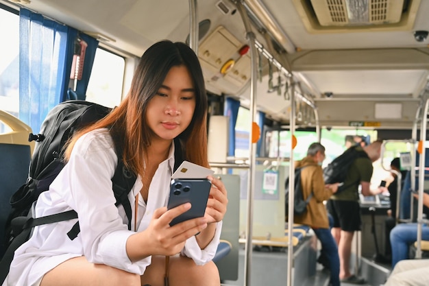 Eine glückliche asiatische Touristin hält eine Kreditkarte und benutzt ein Smartphone, während sie mit dem Bus pendelt