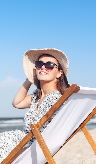 Eine glückliche brünette Frau mit Sonnenbrille und Hut entspannt sich auf einem Holzdeckstuhl am Meerstrand.