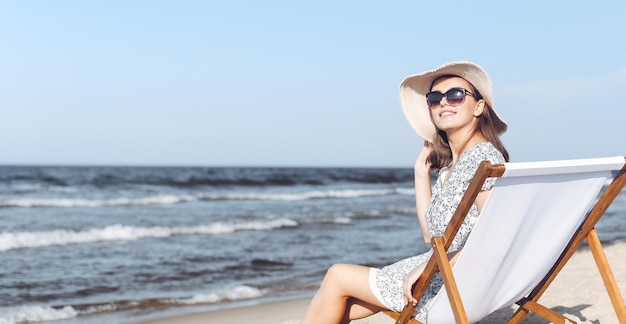 Eine glückliche brünette Frau mit Sonnenbrille und Hut entspannt sich auf einem Holzdeckstuhl am Meerstrand