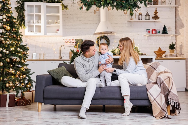 Eine glückliche Familie sitzt mit ihrem kleinen Sohn neben dem Weihnachtsbaum auf der Couch und lächelt freudig Heimfestliche Atmosphäre des neuen Jahres