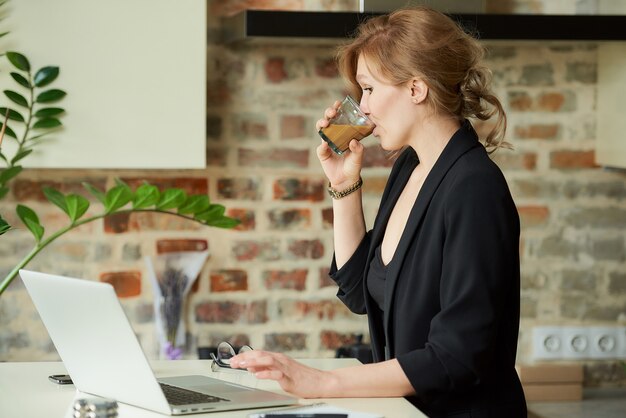 Foto eine glückliche frau, die entfernt an einem laptop in einer küche arbeitet. eine dame, die während einer videokonferenz mit ihren kollegen zu hause einen kaffee trinkt. ein lehrer trinkt, während er sich auf eine online-vorlesung vorbereitet