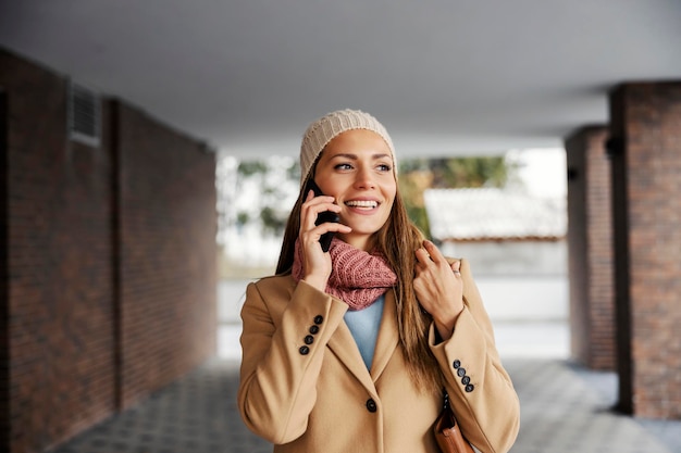 Eine glückliche Frau, die in einem warmen Outfit gekleidet ist, das bei kaltem Wetter im Freien steht und am Telefon spricht Telekommunikation und globales Netzwerk Eine Frau, die draußen am Telefon spricht