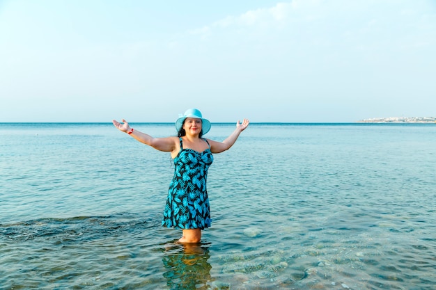 Eine glückliche Frau in einem blauen Sommerkleid und Hut steht im Meer knietief im Wasser.