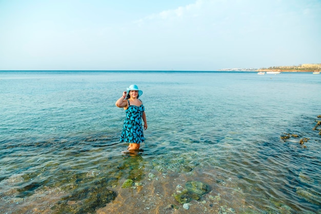 Eine glückliche Frau in einem blauen Sommerkleid und Hut steht knietief im Wasser im Meer. Horizontales Foto.