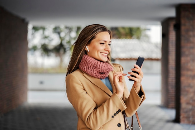 Eine glückliche Frau in einem warmen Outfit, die draußen mit Kopfhörern steht und Musik auf ihrem Telefon auswählt