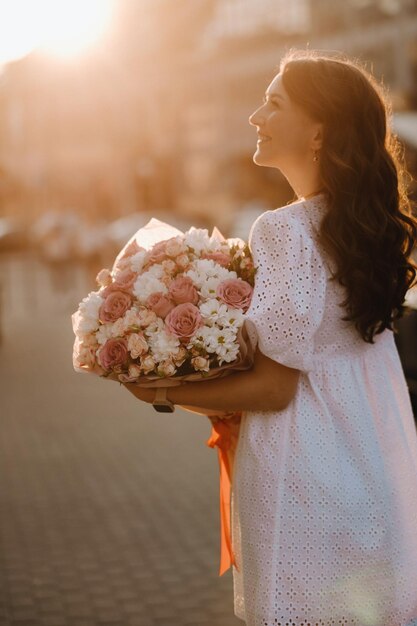 Eine glückliche Frau in einem weißen Kleid bei Sonnenuntergang mit einem Blumenstrauß in der Stadt