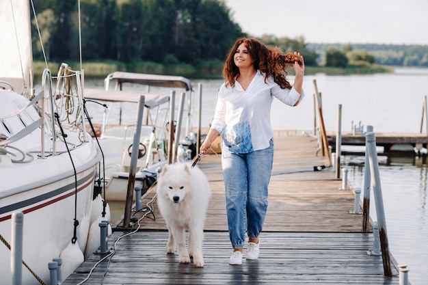 Eine glückliche Frau mit einem großen weißen Hund geht auf dem Pier in der Nähe der Yacht und des Meeres spazieren