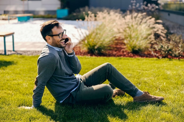 Eine glückliche Geschäftsperson in Smart Casual sitzt auf einem Gras in einem Park und telefoniert