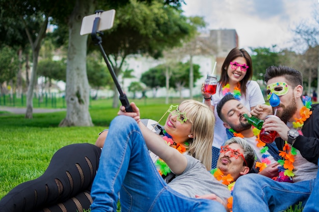 Eine glückliche Gruppe von Freunden, gekleidet in Masken und Geburtstagszubehör, die mit ihrem Selfie-Stick im Gras des Parks Fotos machen.