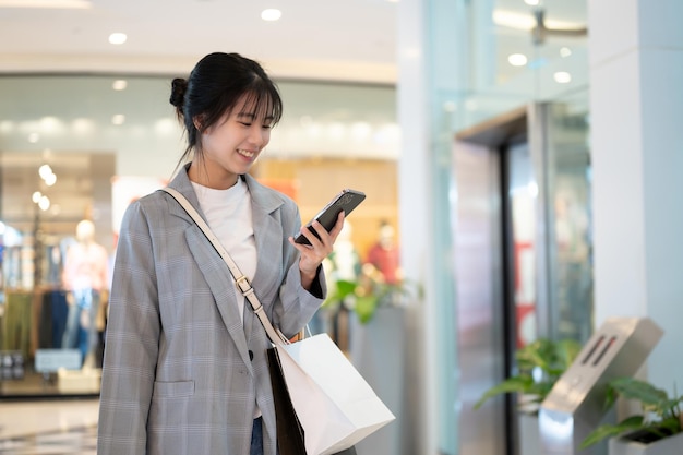 Eine glückliche, hübsche asiatische Frau checkt Nachrichten auf ihrem Handy, während sie durch ein Einkaufszentrum geht
