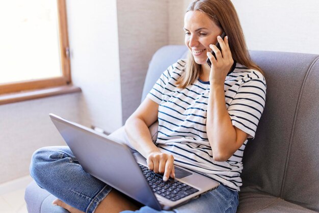 Foto eine glückliche junge frau benutzt ein handy, während sie zu hause mit einem laptop auf dem sofa sitzt eine frau spricht mit einem handy, stellt fragen, schaut auf den computerbildschirm