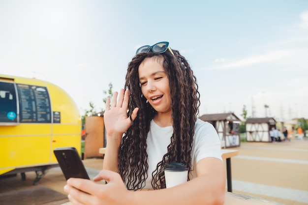 Eine glückliche junge Frau, die über Kopfhörer an einer Webkonferenz teilnimmt, oder ein Freiberufler, der an einem Tisch in einem Café im Freien sitzt und ein Mädchen mit Dreadlocks per Videolink kommuniziert