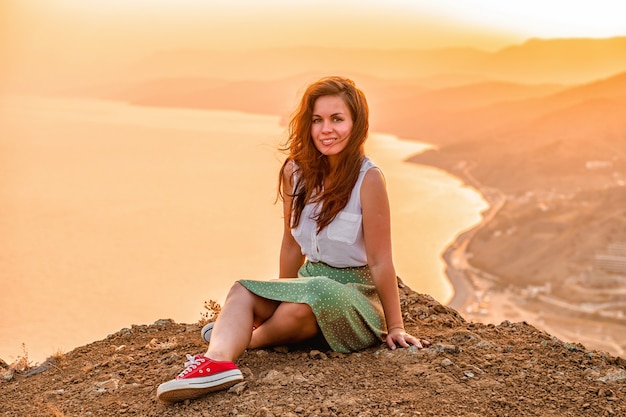 Eine glückliche junge Frau in einem Rock sitzt auf einer Bergklippe unter dem Sonnenuntergangshimmel