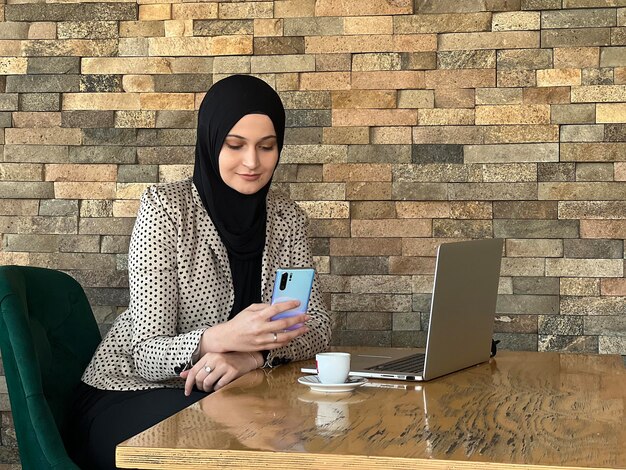 Eine glückliche junge muslimische Frau im Hijab, die an einem Laptop arbeitet und ein Mobiltelefon im Restaurantcafé benutzt