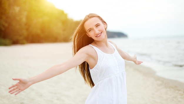 Eine glückliche lächelnde Frau in freier Glückseligkeit steht mit offenen Händen am Meeresstrand. Porträt eines multikulturellen weiblichen Modells in weißem Sommerkleid, das die Natur genießt