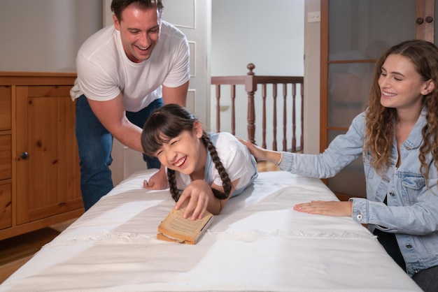 Eine glückliche moderne Familie weckt ihr kleines Mädchen am Wochenende mit spielerischem Kitzeln und drückt damit ihre Liebe und Zuneigung für ihre kleine Tochter aus, die gemeinsam lacht und lächelt