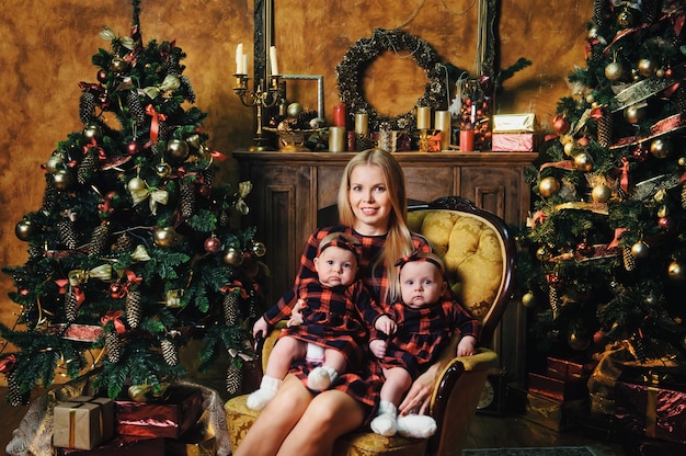 Eine glückliche Mutter mit ihren Zwillingskindern im Neujahrsinneren des Hauses auf dem Hintergrund eines Weihnachtsbaums.