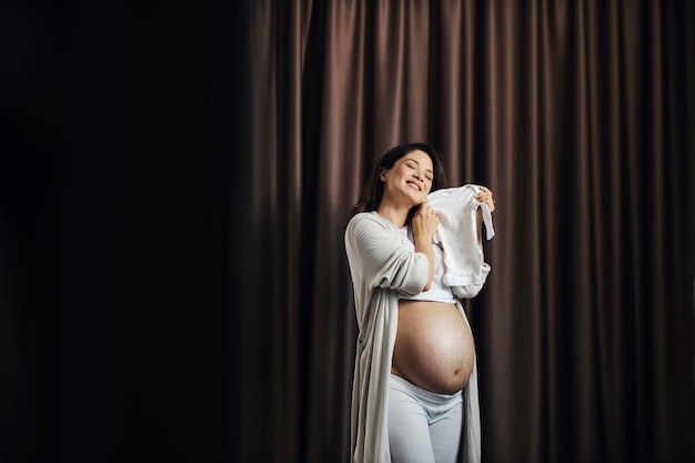 Eine glückliche schwangere Frau steht mit ihrem nackten Bauch mit Dehnungsstreifen und hält Baby-Onsie