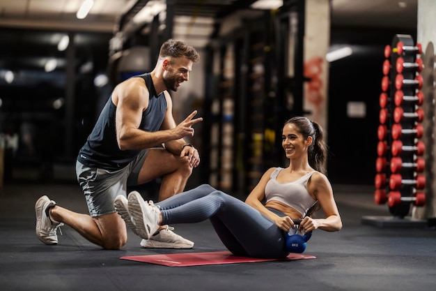 Foto eine glückliche sportlerin macht einen russischen twist mit kesselglocke in einem fitnessstudio und ihr trainer kniet