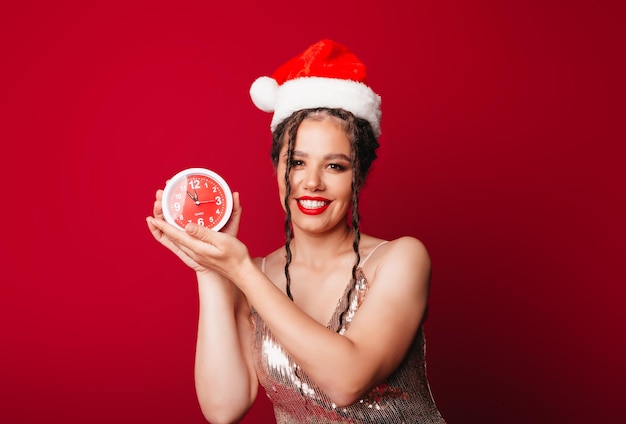 Eine glückliche süße Frau in einem Weihnachtsmann-Hut hält einen Wecker auf rotem Hintergrund Das Konzept der Weihnachtszeit Weihnachten und Neujahr