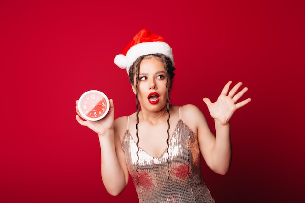 Eine glückliche süße Frau in einem Weihnachtsmann-Hut hält einen Wecker auf rotem Hintergrund Das Konzept der Weihnachtszeit Weihnachten und Neujahr