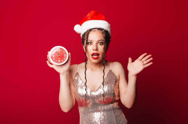 Eine glückliche süße Frau in einem Weihnachtsmann-Hut hält einen Wecker auf rotem Hintergrund Das Konzept der Weihnachtszeit Weihnachten und Neujahr