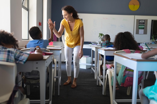 Eine glückliche weiße junge Lehrerin, die einem afroamerikanischen Jungen, der am Schreibtisch sitzt, Highfive gibt