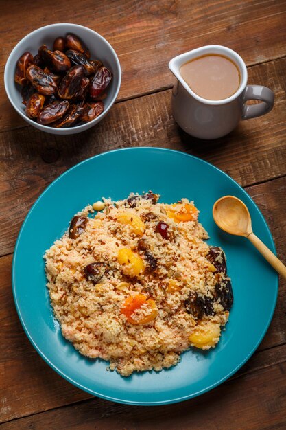 Eine große Schüssel Couscous auf einem Holztisch neben Dattelnüssen und einem Krug mit Fruchtbrühe