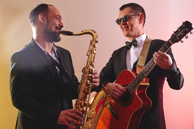 Eine Gruppe aus zwei Musikern, einer männlichen Jazzband, einem Gitarristen und einem Saxophonisten in klassischen Kostümen improvisiert auf Musikinstrumenten in einer Studiobühnenbeleuchtung
